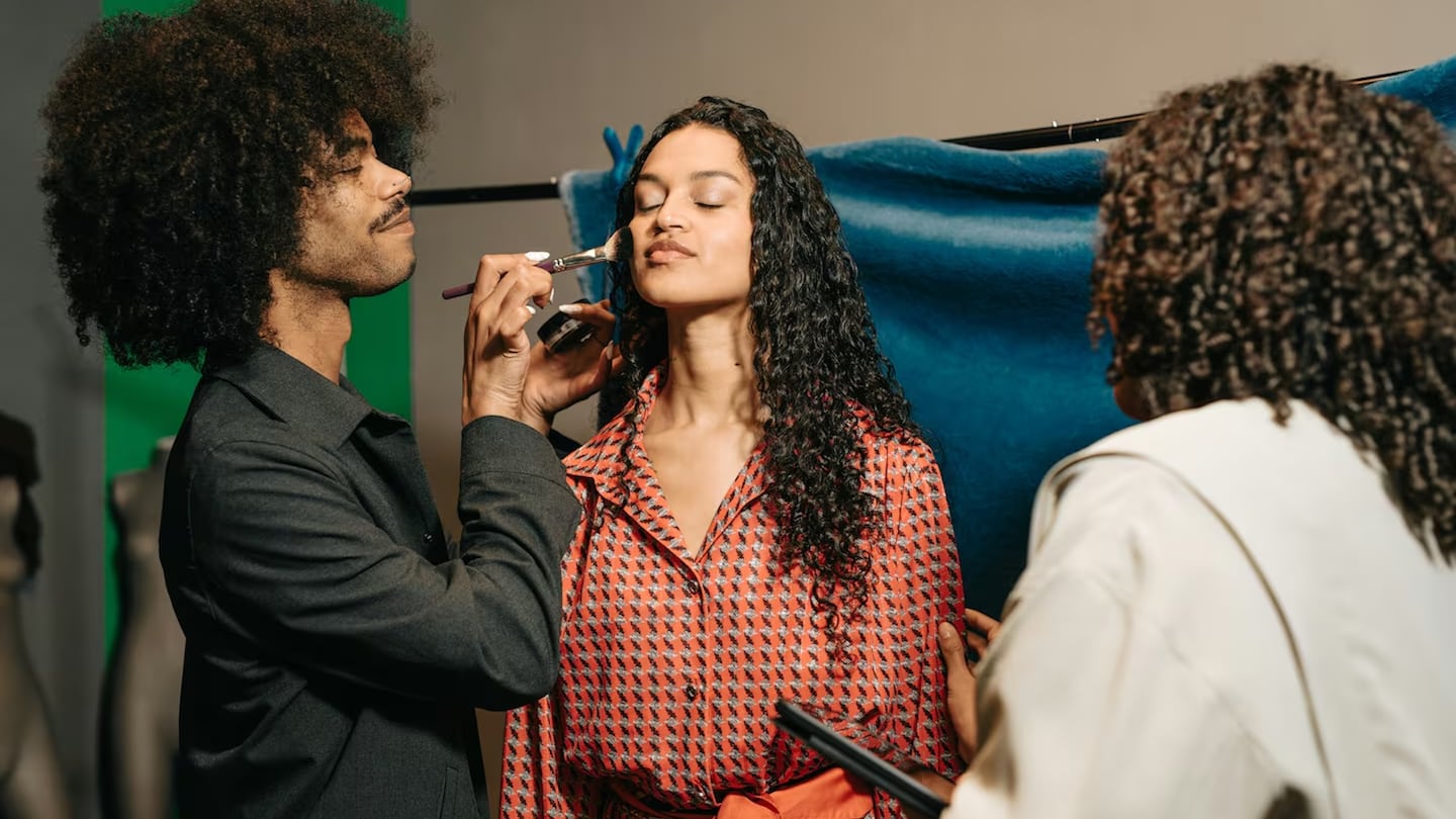 A person with long, dark curly hair and their eyes closed sits for a makeup application. A makeup artist with a large afro and a nose ring stands to the left, using a brush to apply powder to the person's cheek. To the right, another individual with curly hair, seen from behind, assists with the process. The person receiving makeup wears a red and black patterned shirt with a matching belt. The background includes a green screen and a blue fabric backdrop draped over a clothing rack.