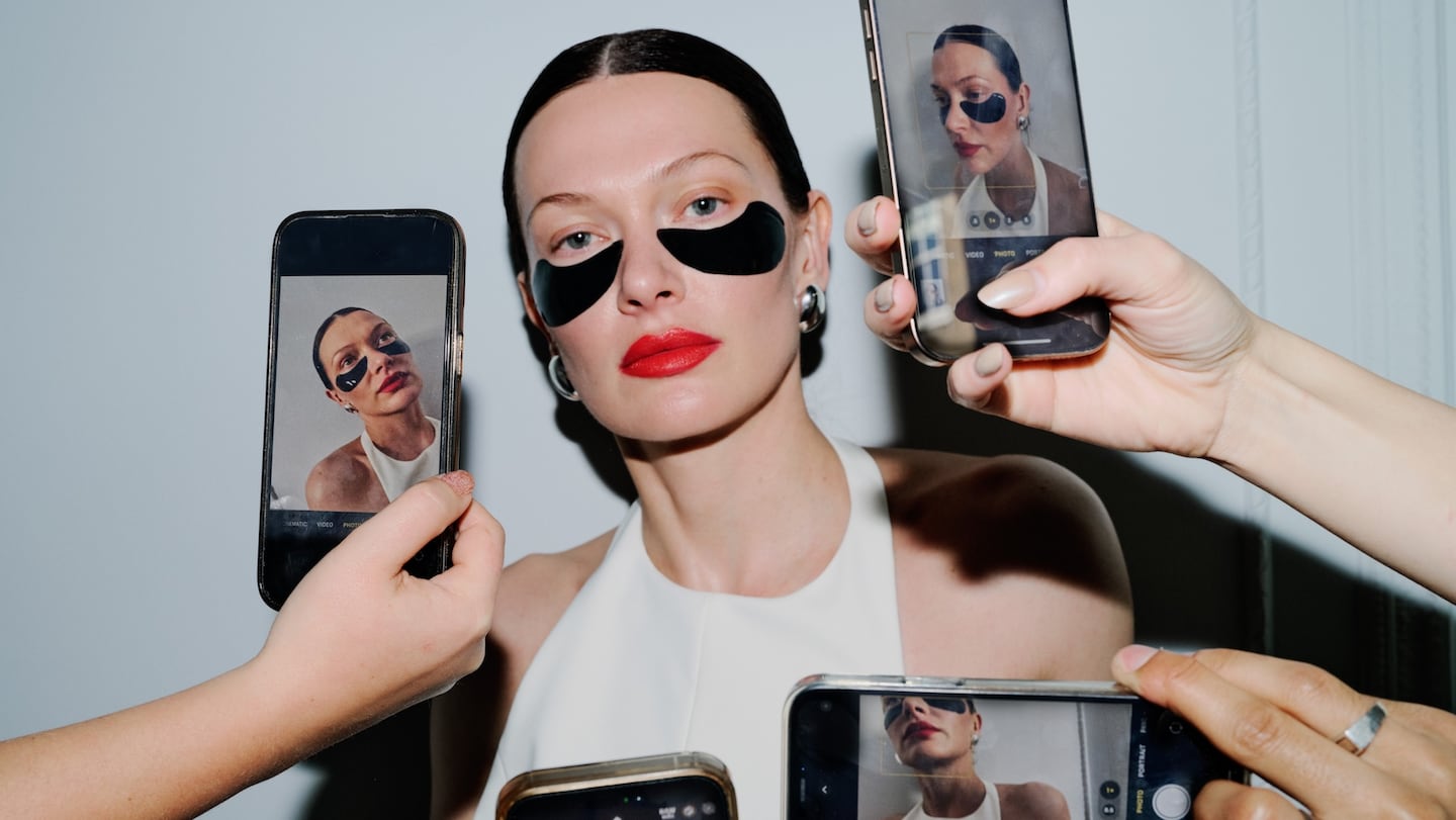 A woman with slicked-back hair and red lipstick wears black under-eye masks while several people hold smartphones taking photos of her from different angles.