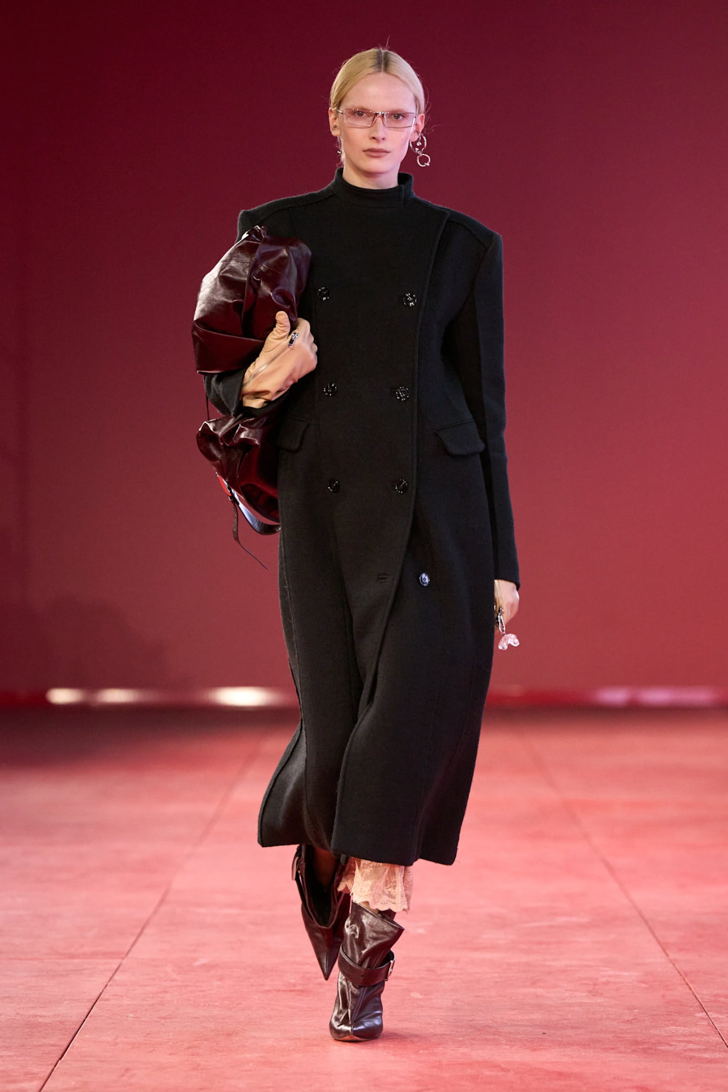 Model on runway wearing long black coat and, shaded sunglasses and a oversized leather clutch.