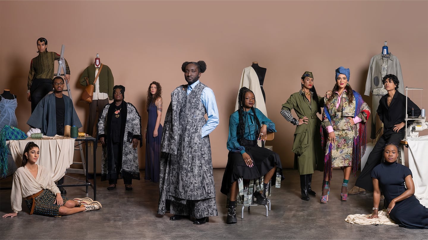 A diverse group of fashion designers from the African Fashion Association (AFA) pose together in a studio setting against a neutral beige backdrop. There are twelve individuals in total, showcasing a wide range of styles, personalities, and expressions. They are arranged in a balanced composition, with some standing, others sitting or perched on props like stools, ladders, or the floor.
The designers wear eclectic outfits that highlight various fashion aesthetics—from structured and tailored garments to fluid, printed, and layered looks. Their clothing features rich textures, bold prints, and thoughtful detailing, reflecting creativity and individuality. Several dress forms and mannequins with garments or accessories are positioned throughout the scene, along with sewing machines, spools of yarn, and fabric, evoking a studio environment that merges craftsmanship and design.
The atmosphere is poised and confident, celebrating cultural identity, inclusivity, and innovation in fashion.